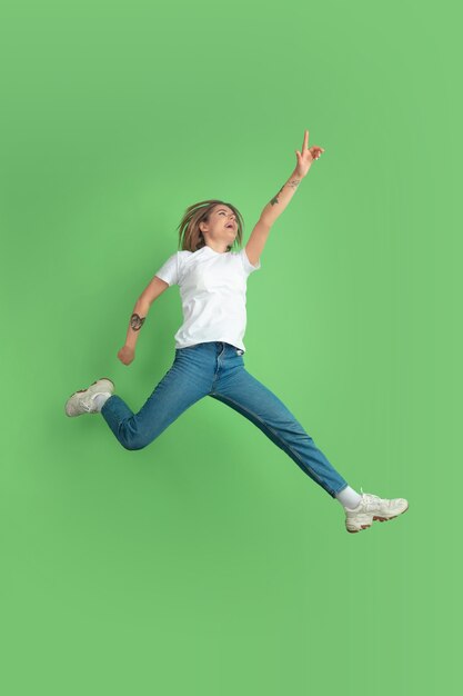 Apuntando hacia arriba en salto. Retrato de mujer joven caucásica aislado en la pared verde. Modelo de mujer hermosa en camisa blanca. Concepto de emociones humanas, expresión facial, juventud.