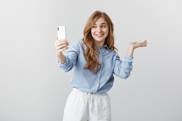 Aquí está mi habitación. Retrato de mujer guapa feliz emocionada en blusa azul mostrando alrededor mientras video chat a través de teléfono inteligente, sonriendo ampliamente, dirigiendo al espacio de copia sobre pared gris