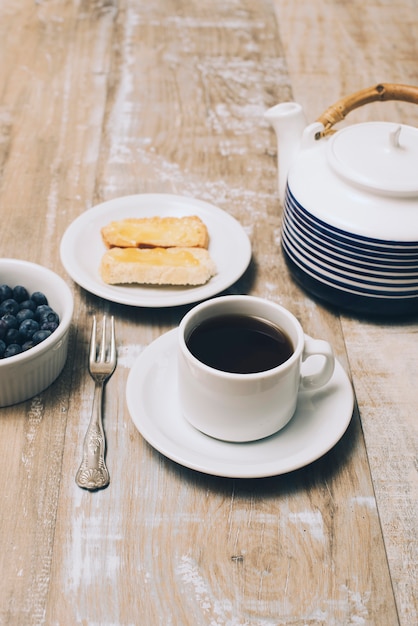 Foto gratuita arándanos; un pan; taza de café y tetera en mesa de madera