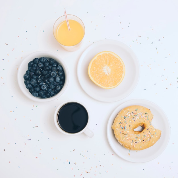 Foto gratuita arándanos; taza de café; naranja a la mitad; jugo y donut con chispitas de colores sobre fondo blanco