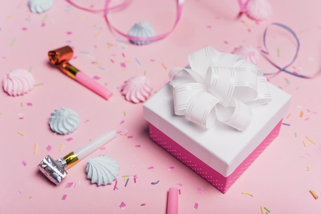Arco de satén blanco en caja de regalo con soplador de fiesta y caramelos sobre fondo rosa