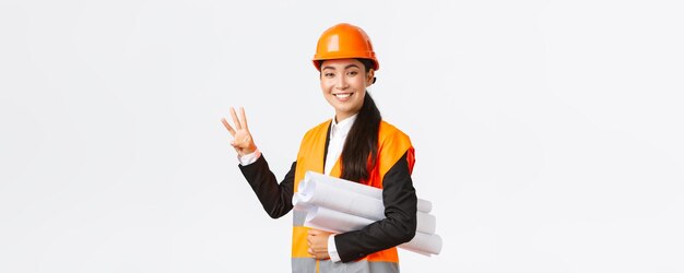 Arquitecta asiática sonriente en el área de construcción que muestra el plano de transporte número tres como si usara casco de seguridad dando tiempo fijo para la tarea realizada fondo blanco