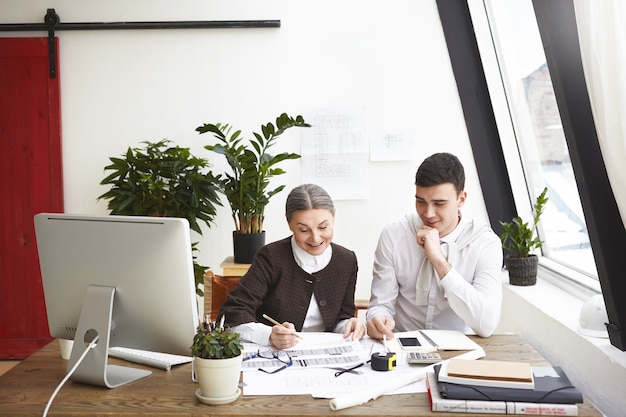 Arquitecta jefa anciana alegre y hombre joven trabajando juntos en un proyecto arquitectónico, felices con los excelentes resultados de su trabajo común, hablando y sonriendo. Trabajo en equipo y colaboración