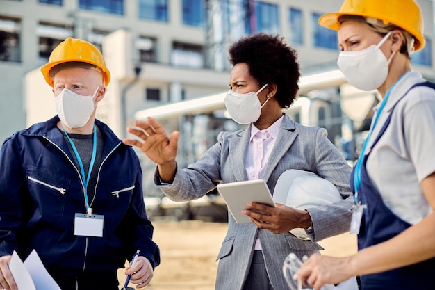 Arquitecta negra e ingenieras civiles con máscaras protectoras hablando en el sitio de construcción