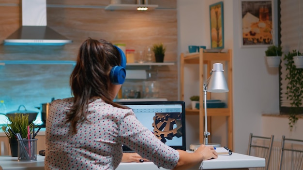 Foto gratuita arquitecto con auriculares inalámbricos usando laptop mientras trabaja en casa por la noche sentado en la cocina. ingeniera industrial que estudia en la computadora personal que muestra el software de cad.