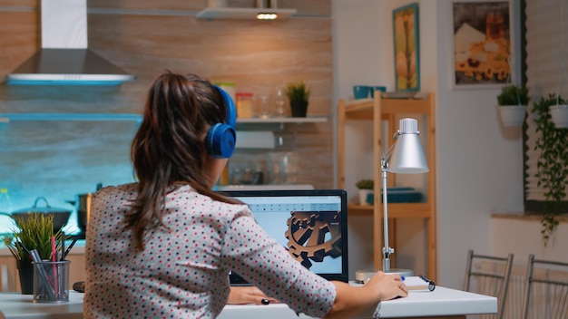 Arquitecto con auriculares inalámbricos usando laptop mientras trabaja en casa por la noche sentado en la cocina. Ingeniera industrial que estudia en la computadora personal que muestra el software de cad.