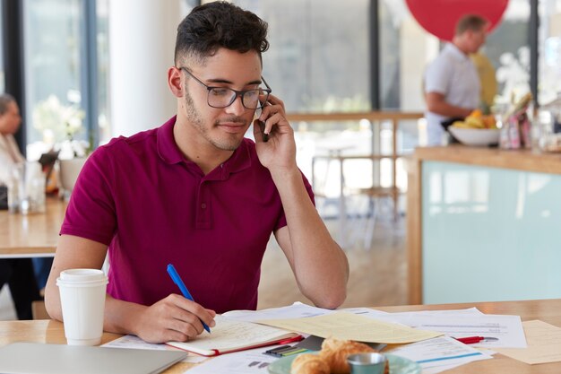 El arquitecto exitoso piensa en la implementación del proyecto, discute ideas con un colega a través del teléfono celular, hace registros en el bloc de notas, disfruta de una bebida fresca en un acogedor bistró. Diseñador masculino trabaja en cafe