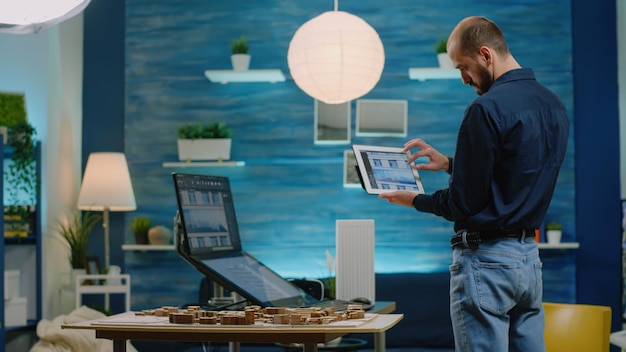 Arquitecto mirando tableta con modelo de construcción para desarrollo. Hombre ingeniero sosteniendo un dispositivo y analizando el diseño de la construcción en la computadora, con maqueta y plan de impresión en la mesa.