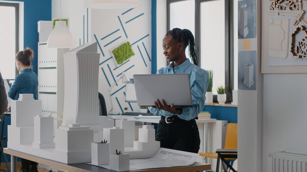 Arquitecto de la mujer que sostiene la computadora portátil para analizar el modelo del edificio en la oficina de arquitectura. Ingeniero trabajando con computadora y maqueta para diseñar estructura de construcción para proyecto urbano.