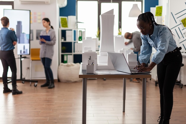 Foto gratuita arquitecto en una oficina de arquitectura moderna que trabaja en una laptop en el escritorio con un modelo de rascacielos de espuma blanca en 3d. ingeniero mirando planos del proyecto de arquitectura del área de desarrollo urbano.