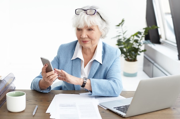 Arquitecto senior de mujer de moda con cabello gris y anteojos en la cabeza navegando por internet o escribiendo mensajes de texto a través de un teléfono inteligente, trabajando en el escritorio de la oficina, sentado frente a una computadora portátil abierta