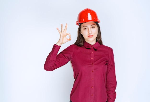 Arquitecto de sexo femenino acertado en pie del casco duro rojo y dando la muestra aceptable.