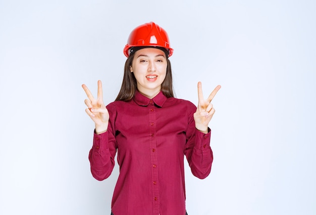 Arquitecto de sexo femenino acertado en rojo casco duro de pie y dando el signo de la victoria.