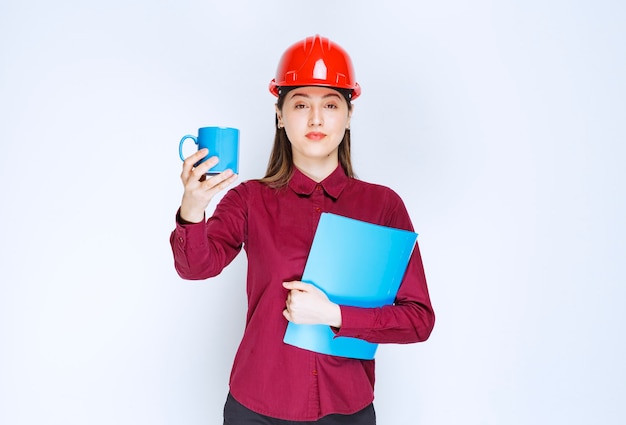 Arquitecto de sexo femenino en documentos del casco rojo en la mano que sostiene el café.