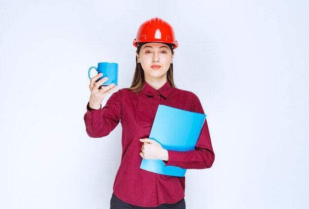 Arquitecto de sexo femenino en documentos del casco rojo en la mano que sostiene el café.