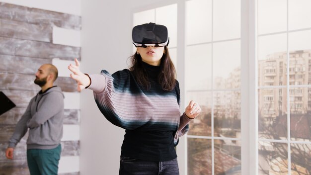 Arquitecto de sexo femenino con gafas de realidad virtual en apartamento vacío y agente de bienes raíces hablando con el cliente en segundo plano.