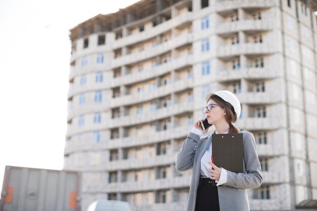 Arquitecto de sexo femenino joven profesional que habla en el teléfono móvil que sostiene el tablero en el emplazamiento de la obra