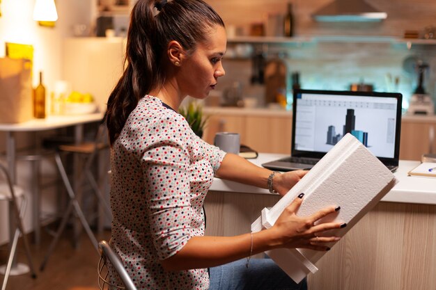 Arquitecto de sexo femenino que mira el modelo de construcción durante la noche en la oficina en casa. Artista ingeniero que crea y trabaja en la oficina con modelo de construcción a escala, determinación, carrera.