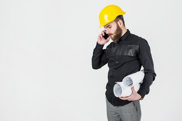 Arquitecto de sexo masculino joven que sostiene impresiones azules que hablan en el teléfono móvil