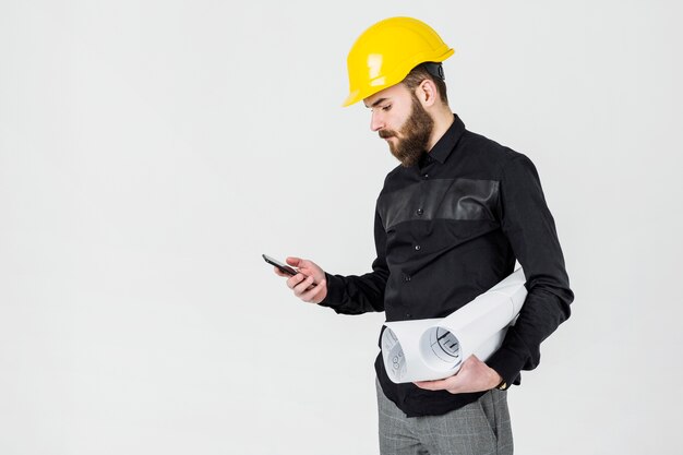 Un arquitecto de sexo masculino que desgasta el casco amarillo que mira el smartphone