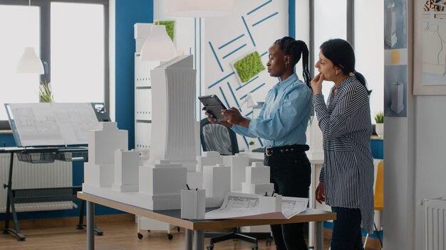 Arquitectos que analizan el modelo de construcción con una tableta digital para trabajar en el diseño de la construcción. Equipo de mujeres diversas que utilizan dispositivos y tecnología para diseñar un plan inmobiliario en una oficina de arquitectura.