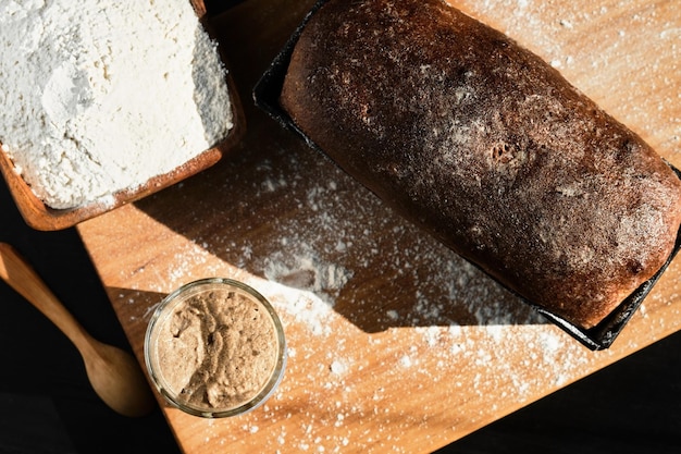 Arrancador activo de masa fermentada de centeno y trigo en un frasco de vidrio junto al ingrediente harina y pan integral recién horneado sobre la mesa