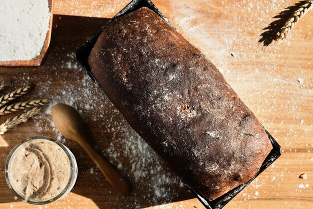 Arrancador activo de masa fermentada de centeno y trigo en un frasco de vidrio junto al ingrediente harina y pan integral recién horneado sobre la mesa