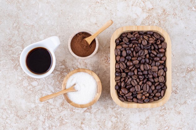 Un arreglo de taza de café, azúcar, polvo de café molido y un montón de granos de café en una bandeja de madera