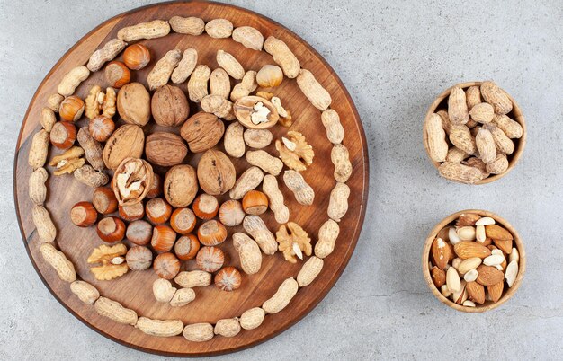 Un arreglo de varios tipos de nueces sobre tabla de madera con cuencos de cacahuetes, almendras y pistachos sobre superficie de mármol.