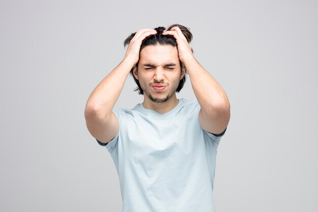 arrepentido joven guapo manteniendo las manos en la cabeza con los ojos cerrados aislado sobre fondo blanco.