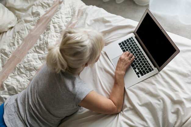 De arriba mujer mayor que usa la computadora portátil en la cama
