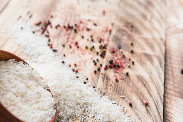 Arroz blanco de grano que se derrama fuera del tazón de madera redondo con; Pimientos negros frescos sobre superficie texturizada de madera