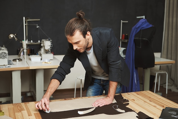 Artista creando obra maestra. Cerca del diseñador de ropa masculina hermosa joven con peinado de moda y ropa de moda trabajando en vestido nuevo con expresión de la cara concentrada.