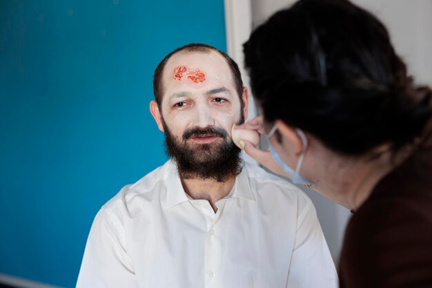 Artista dibujando una mirada de zombi en el hombre, aplicando un maquillaje sangriento y dramático con un disfraz de terror no muerto. Usando efectos especiales para crear una cara de cadáver terrible y espeluznante con ojos locos.