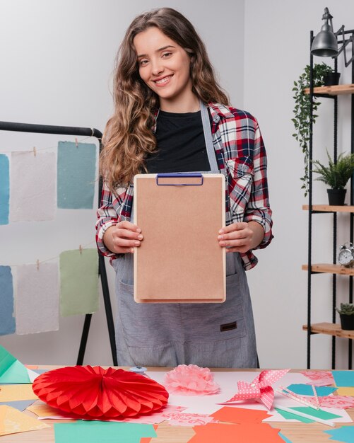 Artista femenina atractiva feliz mostrando portapapeles con papel marrón liso