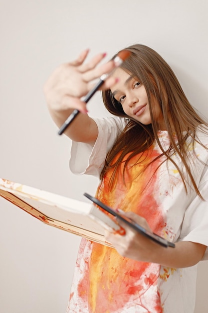 Artista femenina de pie cerca de una pared blanca sosteniendo un lienzo y pinceles