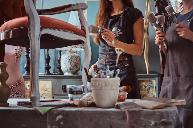 Artista femenina con su asistente en delantales pintando silla vintage con pincel en taller.