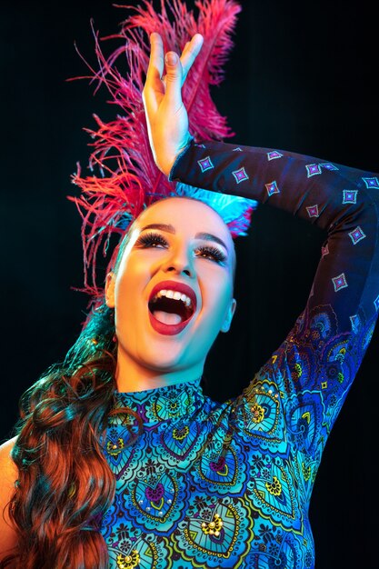 Artista. Hermosa mujer joven en carnaval, elegante disfraz de mascarada con plumas sobre fondo negro en luz de neón. Copyspace para anuncio. Celebración de fiestas, baile, moda. Tiempo festivo, fiesta.