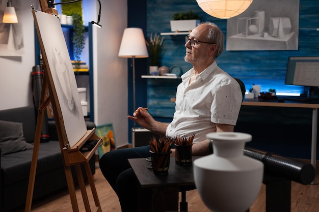 Artista jubilado sosteniendo obras de arte de dibujo a lápiz admirando el progreso en la noche sentado en el estudio de casa. Retrato de un anciano creador de arte frente a un caballete con un boceto haciendo bocetos creativos.