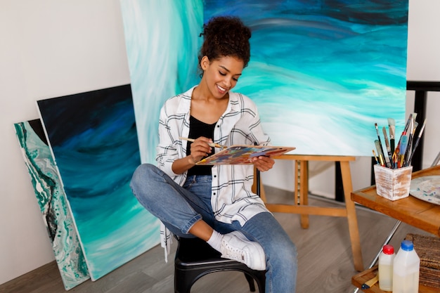 Artista de mujer negra en estudio sosteniendo un pincel. Estudiante inspirado sentado sobre sus obras de arte.