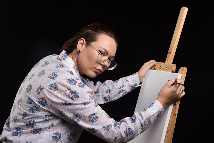 Foto gratuito artista mujer con pincel y pinturas en sus manos se encuentra cerca del caballete y mira a la cámara. foto de alta calidad