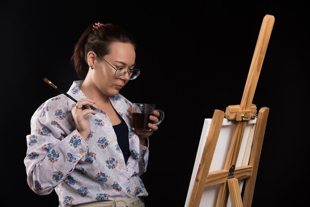 Artista mujer sosteniendo pincel y mirando lienzo sobre fondo negro