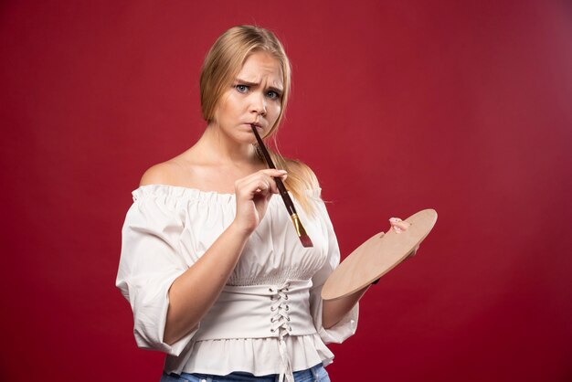 La artista rubia sostiene una paleta y pinceles y se ve sorprendida y confundida.