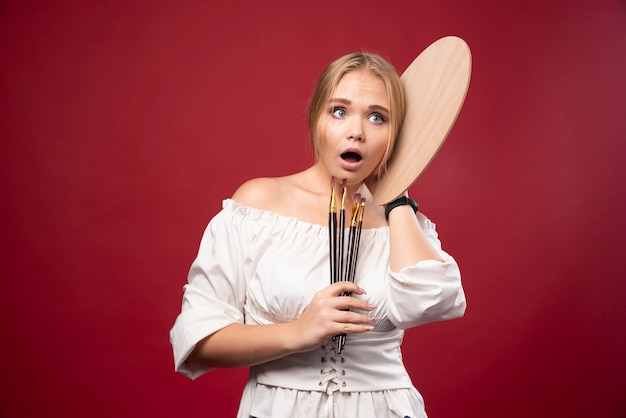 La artista rubia sostiene una paleta y pinceles y se ve sorprendida y confundida.