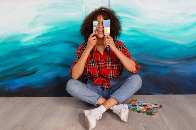 Artista sonriente de la mujer negra en el estudio que sostiene un cepillo. Estudiante inspirado sentado sobre sus obras de arte.