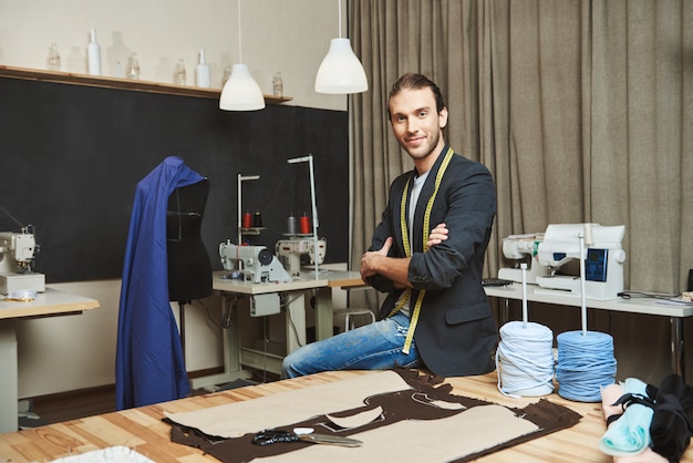 Artista y su lugar de trabajo. Retrato del diseñador de ropa masculino caucásico atractivo maduro con elegante peinado y traje de moda sentado en su estudio, posando para la foto en la revista de moda.