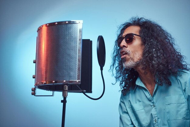 Artista vocal africano con una camisa azul escribiendo emocionalmente una canción en el estudio de grabación. Aislado sobre un fondo azul.