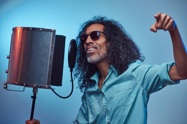 Artista vocal africano con una camisa azul escribiendo emocionalmente una canción en el estudio de grabación. Aislado sobre un fondo azul.