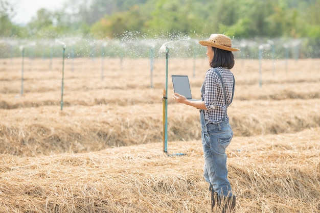 Asia joven agricultora con sombrero de pie en el campo y escribiendo en el teclado de la computadora portátil. Mujer con laptop supervisando el trabajo en tierras de cultivo, concepto de ecología, transporte, aire limpio, alimentos, productos biológicos