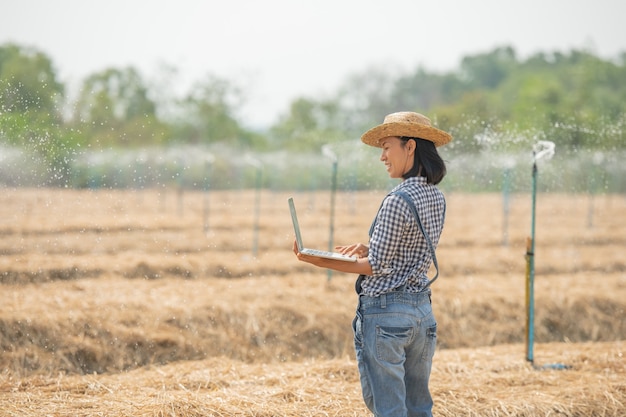 Asia joven agricultora con sombrero de pie en el campo y escribiendo en el teclado de la computadora portátil. Mujer con laptop supervisando el trabajo en tierras de cultivo, concepto de ecología, transporte, aire limpio, alimentos, productos biológicos